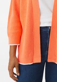 Korallen-orange Strickjacke mit Rippdetails, offenem Frontbereich und kurzen Ärmeln; kombiniert mit dunkelblauen Jeans.