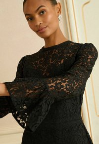 Fekete csipkebársony ruha hosszú, bővülő ujjakkal és testhezálló felsőrészsel, virágmintákkal díszítve és fodros dekoltázzsal.