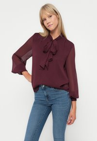 Blusa bordeaux con dettaglio al collo legato e maniche trasparenti e fluide, abbinata a jeans skinny blu su uno sfondo bianco semplice.