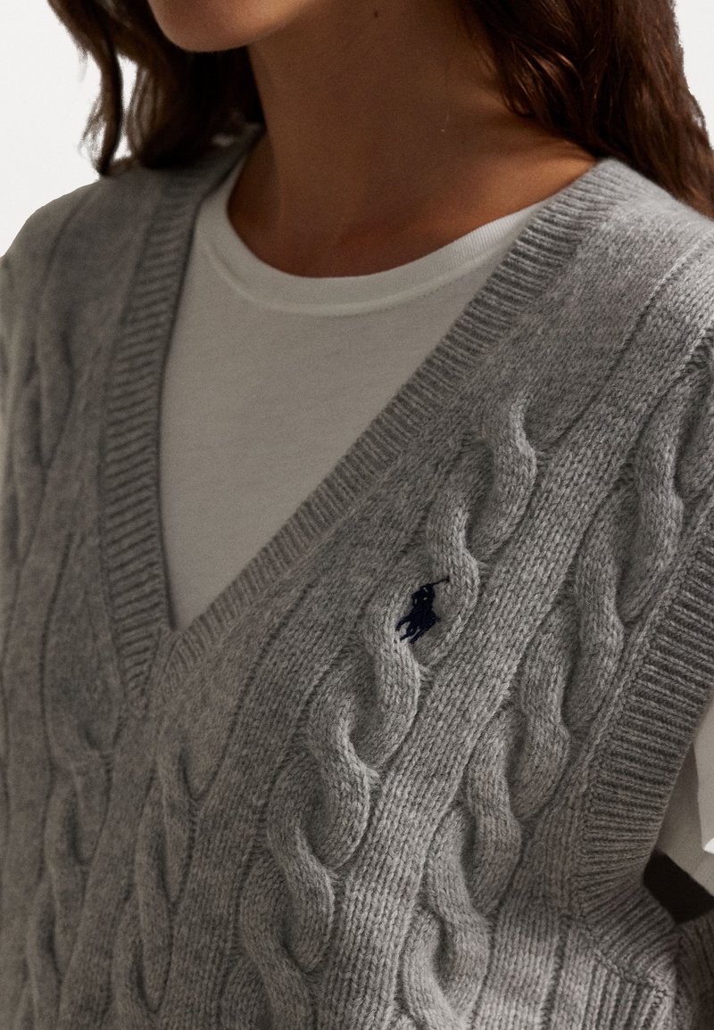 Grå kabelstrik sweater vest med V-hals design og ribbede kanter, prydet med et lille marineblåt logo på forsiden.