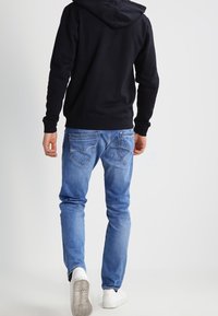 Hombre vestido con una sudadera negra, jeans claros y zapatillas blancas, de pie de espaldas contra un fondo liso claro.