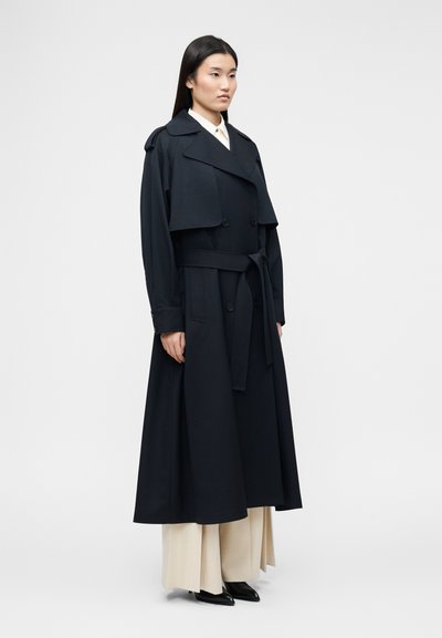 Femme portant un long trench-coat noir à double boutonnage avec ceinture, par-dessus un pantalon beige à jambes larges et des chaussures noires pointues, debout de profil.