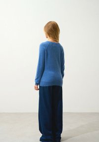 Blå tröja med en slät textur, ribbade muddar och fåll, i kombination med vida mörk denimbyxor. Bakvyn mot en enfärgad bakgrund.