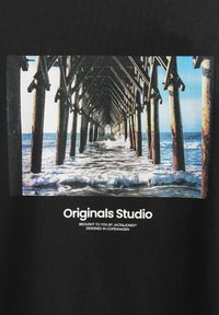 Juodas audinys su stačiakampiu grafiku, vaizduojančiu prieplauką virš vandens, su medinėmis atramomis ir bangomis, apačioje užrašyta "Originals Studio".
