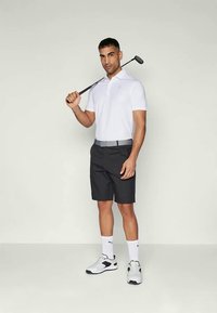 Golfový outfit skládající se z bílé polo košile, černých šortek, šedého pásku a černobílých golfových bot. Hráč drží golfovou hůl.