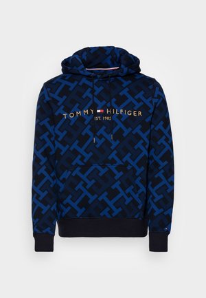 Blau-schwarzer Kapuzenpullover mit Muster, Fronttasche, Kordelzug und dem Schriftzug "Tommy Hilfiger EST. 1985" mit kleinem Flaggenlogo auf der Brust.