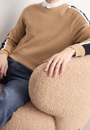 Persoon wearing een tan gebreide trui met zwart-witte gestreepte mouwen, blauwe spijkerbroek en een zilveren ring, hand rustend op een beige kussen met textuur.