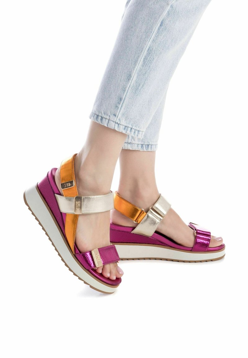 Pieds portant des sandales à plateforme colorées avec des brides roses, orange et dorées, associées à un jean bleu clair retroussé sur fond blanc.