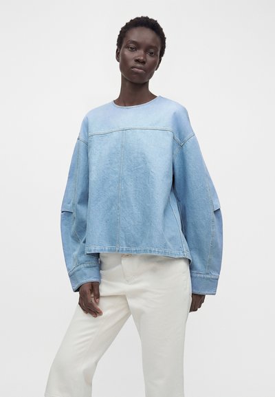 Jeune personne portant une chemise en jean bleu clair oversize à manches longues et un pantalon blanc, debout devant un fond clair uni.