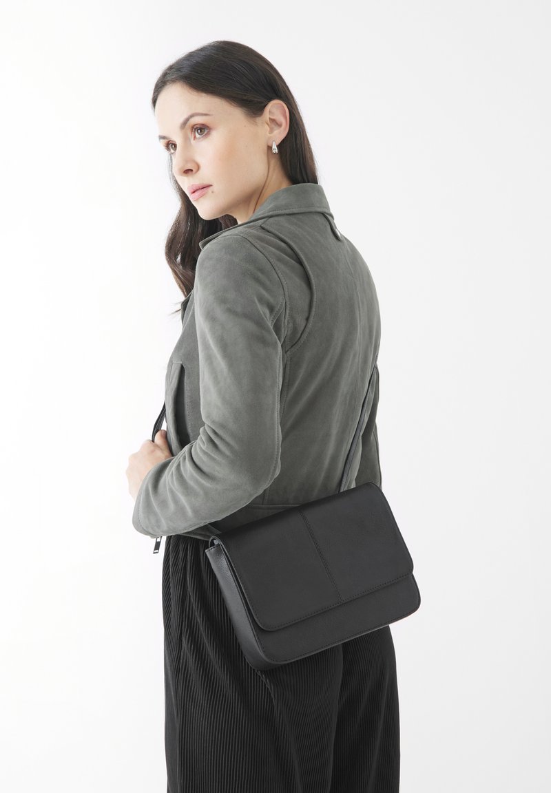 Bolso cruzado de cuero negro con cierre de solapa, textura suave y diseño minimalista. Llevado sobre el hombro con una chaqueta verde olivo.