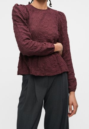 Femme portant un chemisier bordeaux texturé à manches longues avec des épaules bouffantes et un pantalon plissé gris foncé, debout avec un bras croisé sur son corps.
