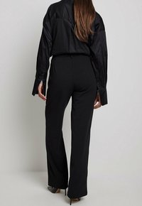 Schwarzes Langarmshirt mit drapiertem Rücken, kombiniert mit hoch taillierten schwarzen Hosen und spitzen schwarzen High Heels. Glatte, formelle Stoffoberfläche.