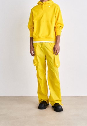 Persona con sudadera con capucha amarilla brillante y pantalones cargo, calzando zapatos negros gruesos, de pie contra una pared blanca lisa y suelo beige.