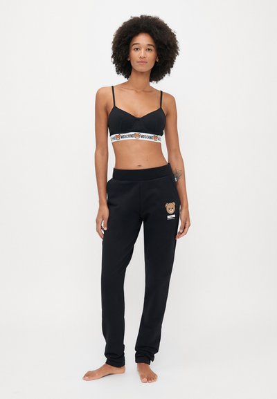 Melns bralette ar baltu Moschino logo joslu, apvienots ar melniem sporta biksēm ar lāča logo, abi izgatavoti no mīkstas, elastīgas auduma.