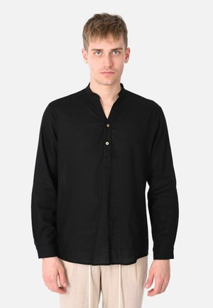 Giovane uomo che indossa una camicia nera a maniche lunghe con colletto a V abbottonato e pantaloni beige, che posa davanti a uno sfondo chiaro e semplice.