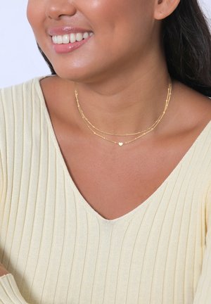 Gold layered necklaces with a heart charm, featuring delicate chain designs and intricate textures, worn with a light-colored ribbed top.