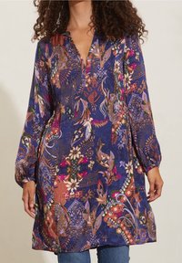 Šaty s květinovým potiskem v odstínech modré, fialové a růžové; mají výstřih do V, dlouhé balonové rukávy a splývavou siluetu.