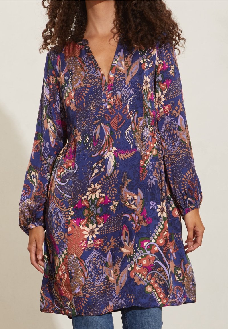 Šaty s květinovým potiskem v odstínech modré, fialové a růžové; mají výstřih do V, dlouhé balonové rukávy a splývavou siluetu.