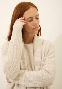 Cárdigan de punto color beige claro con un patrón texturizado, de apertura delantera y mangas largas, superpuesto a un suéter a juego. Tejido suave y cálido.