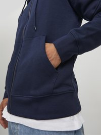Sudadera con cremallera en color azul marino, con un bolsillo frontal, tela texturizada, puños y dobladillo acanalados; llevada sobre una camisa blanca.