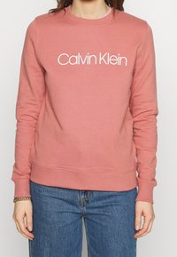 Sudadera de algodón rosa con un logotipo blanco de "Calvin Klein" en la parte frontal, cuello redondo y puños y dobladillo acanalados. Combinada con jeans azules.