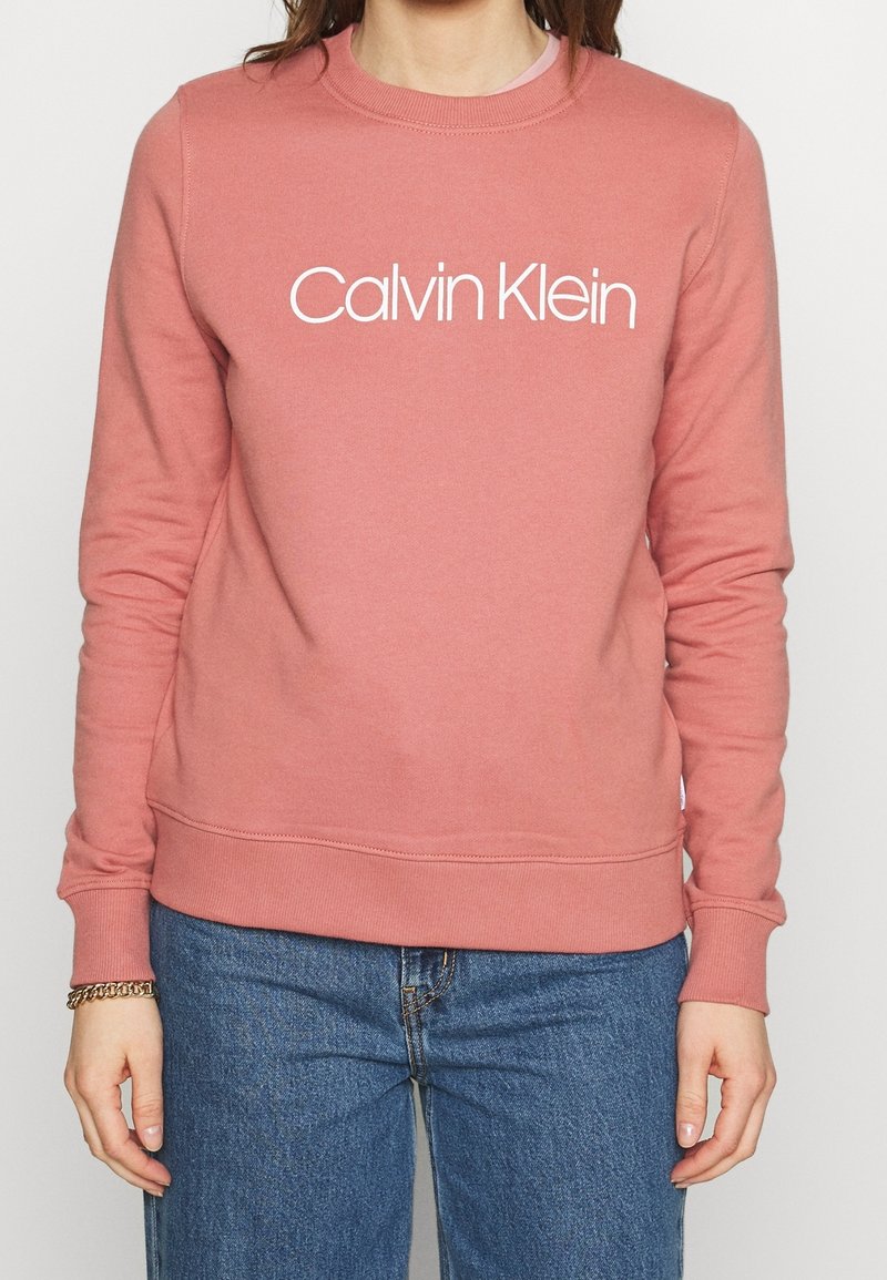 Sudadera de algodón rosa con un logotipo blanco de "Calvin Klein" en la parte frontal, cuello redondo y puños y dobladillo acanalados. Combinada con jeans azules.