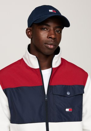 Chaqueta de cremallera roja y marino con cuerpo de forro polar blanco, que cuenta con un bolsillo en el pecho y el logotipo de "Tommy Jeans". Se lleva con una gorra marina.