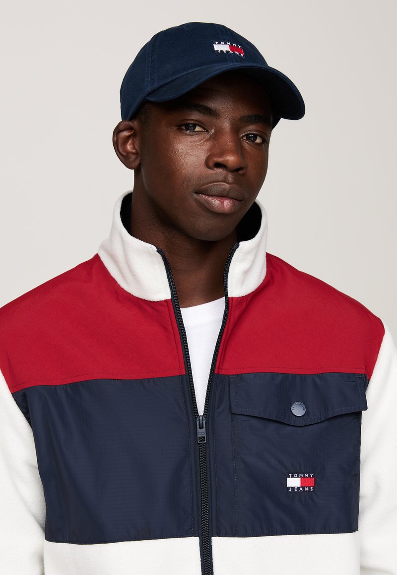 Jachetă cu fermoar roșie și bleumarin, cu corp din fleece alb, având un buzunar pe piept și logo-ul "Tommy Jeans". Purtați cu o șapcă bleumarin.