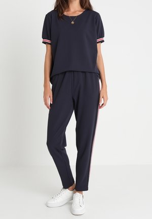 Kvinde iført navyblå kortærmet top og matchende bukser med sidelister, stylet med hvide sneakers og et guldvedhængs halskæde.