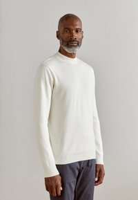 Pull crème, à manches longues, avec un col et des poignets côtelés, associé à un pantalon foncé ; texture lisse et design simple.