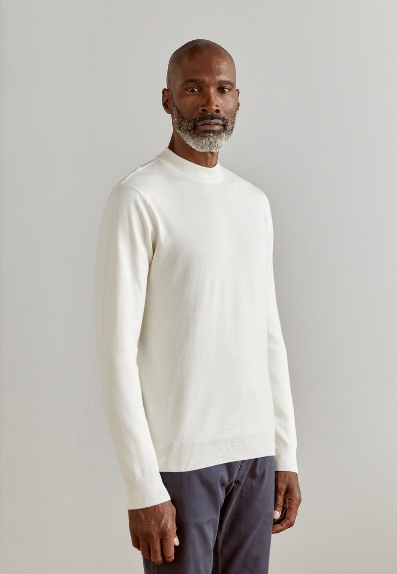 Pull crème, à manches longues, avec un col et des poignets côtelés, associé à un pantalon foncé ; texture lisse et design simple.