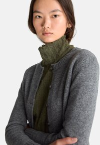 Šedý vlněný cardigan s kulatým výstřihem a knoflíky. Pod ním je částečně vidět zelený texturovaný top s vysokým výstřihem.