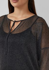 Schwarzes transparentes Oberteil mit strukturierter Oberfläche, das einen Schlitz am Ausschnitt und dekorative Punkte aufweist. Weicher Stoff und eine fließende Silhouette.