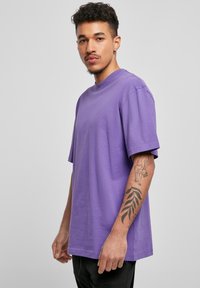 Tricou din bumbac purpuriu, cu decolteu rotund și mâneci scurte, având o croială lejeră. Pantaloni negri și un tatuaj vizibil pe braț.