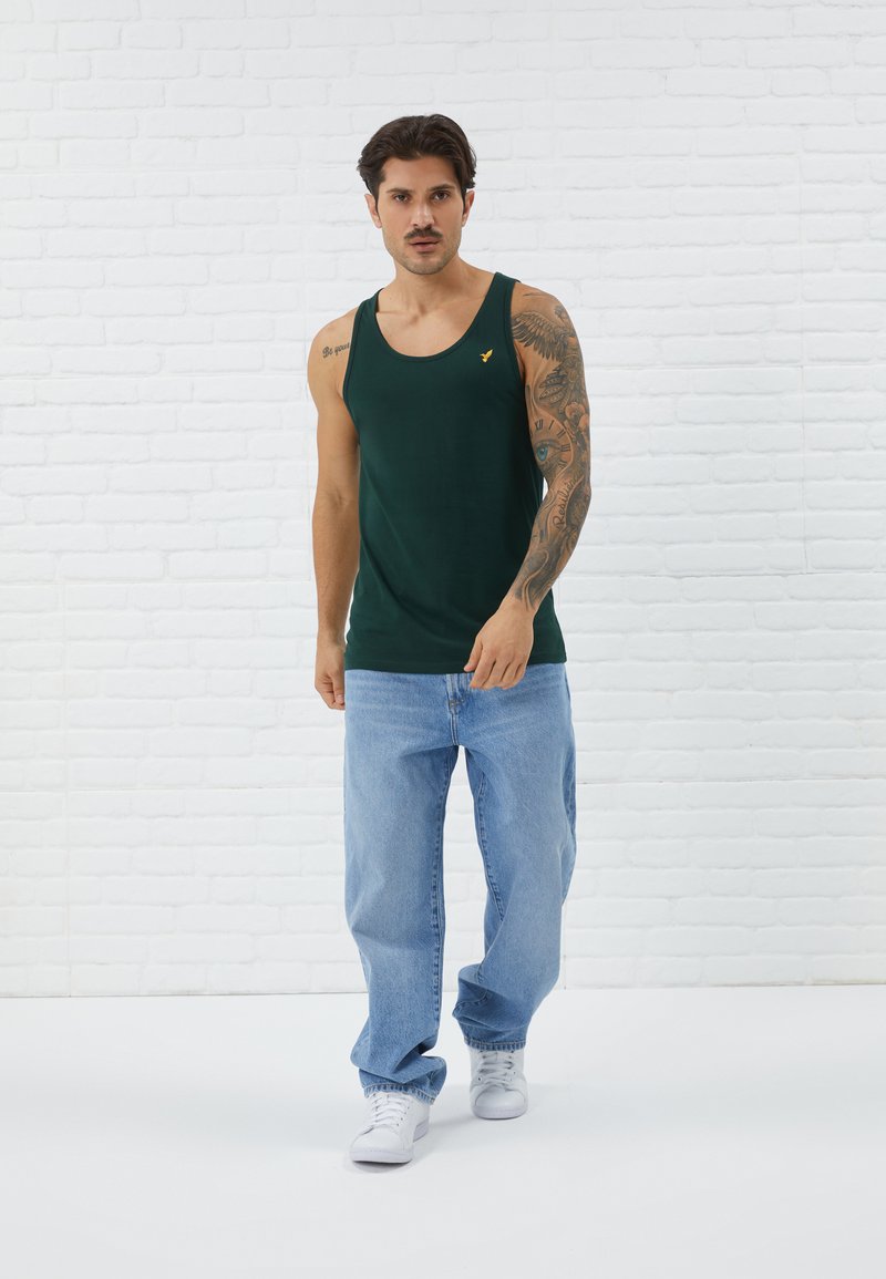 Donkergroen tanktop met een klein geel logo, gecombineerd met lichtblauwe baggy jeans en witte sneakers, tegen een achtergrond van witte bakstenen.