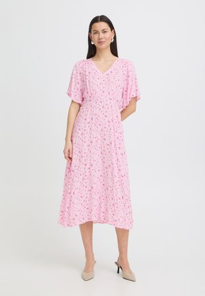 Hosszú, sötét hajú nő, aki rózsaszín virágos midi ruhát visel fodros ujjakkal és bézs magassarkú cipőben áll, egyszerű háttér előtt.