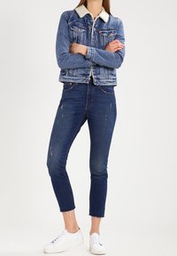 Giacca di jeans con colletto bianco in pelliccia sintetica, abbinata a jeans blu strappati e sneakers bianche. Presenta un taglio corto e un orlo sfrangiato.