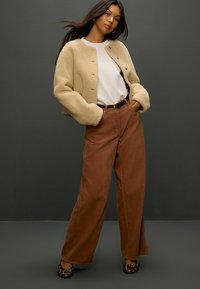 Beige Fleece-Bomberjacke mit Rundhalsausschnitt, fünf Knöpfen, kombiniert mit hoch taillierten braunen Cordhosen und schwarzen gemusterten Schuhen.