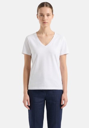 Giovane donna con i capelli raccolti, che indossa una maglietta bianca a scollo a V con maniche corte e pantaloni blu scuro, in piedi davanti a uno sfondo semplice.