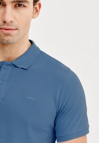 Jasnoniebieska koszulka polo z kołnierzykiem, dwoma guzikami i krótkimi rękawami. Posiada subtelne logo "P L" na lewej części klatki piersiowej.