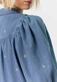 Veste en denim bleu avec des manches bouffantes, ornée de petites broderies florales blanches et jaunes dans le dos et sur les épaules. Texture douce.