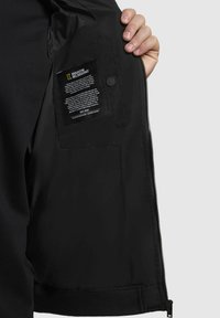 Schwarze Jacke mit einer glatten, matten Oberfläche; verfügt über eine sichtbare Innentasche mit Pflegeetikett und einen Reißverschluss auf der Vorderseite.