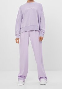 Lichtpaars sweatshirt met omgeslagen mouwen, voorzien van een witte scriptlogo. Gecombineerd met losse lichtpaarse broek en witte sneakers.