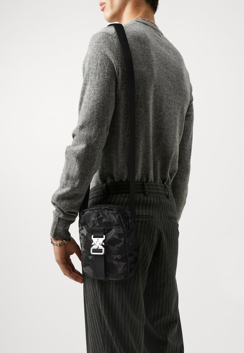 Borsa a tracolla camouflage nera con hardware metallico, tracolla regolabile e finitura in tessuto morbido. Caratterizzata da una chiusura zip sicura e da una forma compatta.