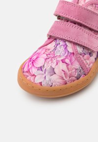 Różowe dziecięce buty w kwiatowy wzór z dwiema paskami na rzepy, z zamszowej górnej części, tanową gumową podeszwą oraz widocznym przeszyciem wzdłuż krawędzi.