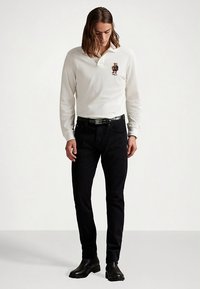 Hvit langermet poloshirt med krage og brodert bjørnelogo. Kombinert med svarte jeans og svarte ankelstøvler. Jevn tekstur.