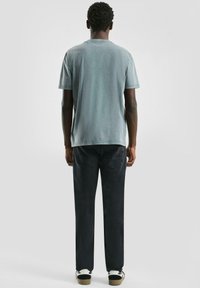 Mężczyzna stojący tyłem, ubrany w wyblakły niebieski t-shirt, czarne dżinsy i czarno-białe sneakersy, na jednolitym szarym tle.