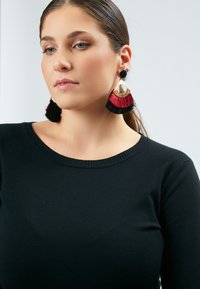 Orecchini con frange rossi e neri con dettagli dorati, caratterizzati da un elemento metallico circolare e texture morbide, indossati con un maglione nero a maglia.