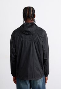 Veste noire légère avec capuche, présentant une texture lisse et des poignets élastiques. Le dos a un design minimaliste avec une couture horizontale.