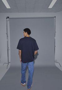 Muž s kudrnatými vlasy, oblečený v černém tričku, modrých džínách a teniskách, stojí čelem k šedému pozadí v ateliéru.